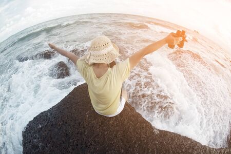 Asian pretty female sitting and resting on rocks with strong waves and holding a Binoculars.の写真素材