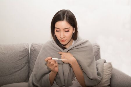 Asian woman sick from cold and used with thermometer suffering on sofa in the room.の写真素材