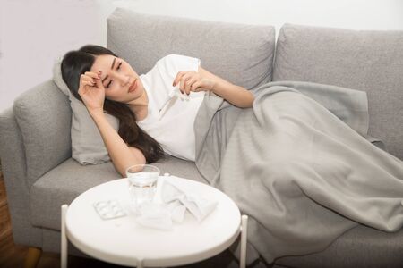 Pretty Asian woman sick from cold and used with thermometer lying on sofa in the room.の写真素材