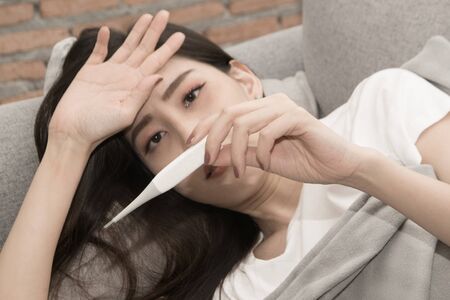Pretty Asian woman sick from cold and used with thermometer lying on sofa in the room.の写真素材