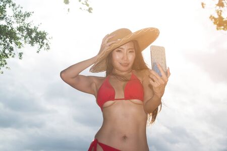 An Asian woman in a mobile-phone swimsuit poses herself, she is a beautiful Asian traveler in a sexy bikini, smiling, taking a selfie with a smartphone on the beach.の写真素材