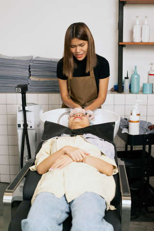 A hairdresser washes hair for a customer in a barber shop and women lying on a chair in a robe which takes care of the hair and scalp professionally.の写真素材
