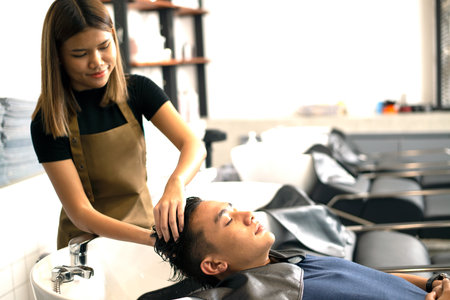 A hairdresser washes hair for a customer in a barber shop and male lying on a chair in a robe which...