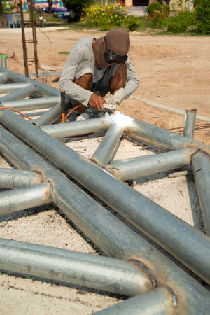 A welder is sitting on a steel roof frame and is welding steel pipes and wearing masks and leather...