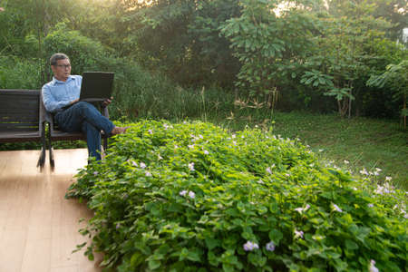 An elderly Asian man working on a laptop sitting in a chair outside the house in the morning. remote...