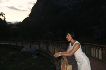 Portrait photography of a mature, beautiful, happy Asian woman. Model in white casual clothes. Standing and smiling at the camera, standing on a bridge with mountains behind.の写真素材