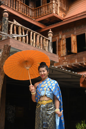 Beautiful Asian woman in Thai National costume holds a paper umbrella posing for photos at a Thai house.の写真素材