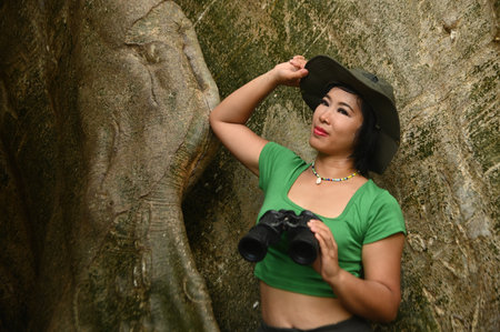 Pretty Asian woman Holding binoculars at the roots of a large tree and are delighted happily. This tree is very large and is a popular attraction for Thai tourists.の写真素材