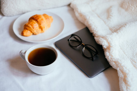 Big white morning cup of coffee and crossant in bed with notebook, glasses, pillow and cosy blanket.の写真素材