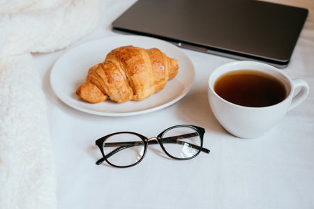 Big white morning cup of coffee and crossant in bed with notebook, glasses, pillow and cosy blanket.の写真素材