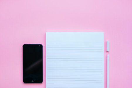 Beautiful office stationery flatlay with ruled notebook, white pen and smartphone on the bright desk with pink background.の写真素材
