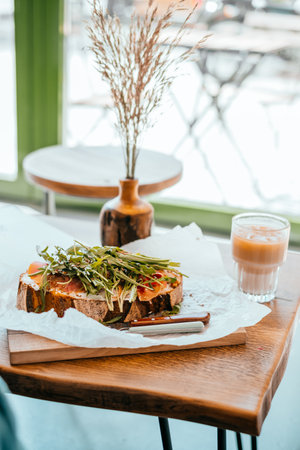 Cup of beautiful cold ice latte and toast with prosciutto and ruccola with wooden vase distichlis spicata on the wooden table. Cafe flatlay situation.の写真素材