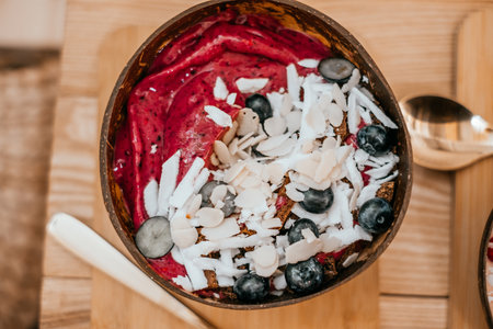 Beautiful ruby berry smoothie bowl with banana, raspberries, blueberries, coconut shavings, chocolate and almond flakes on the wooden table with gold spoon.の写真素材