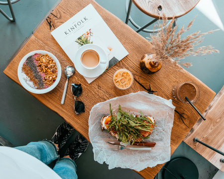 Kyiv, Ukraine - April 29, 2018: Breakfast at cafe. Cup of cold ice latte, toast with prosciutto and ruccola, smoothie bowl with cutlery, on the table with and the issue of Kinfolk magazine .のeditorial素材