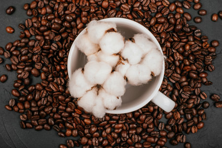 White cup with three cotton flowers and roasted brown coffee beans on the black concrete stone background. Flatlay style.の写真素材