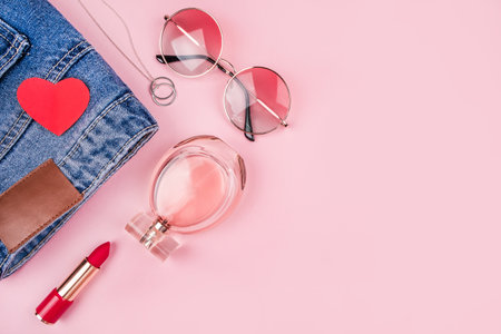 Valentines day fashion and beauty flatlay with blue mom jeans, bottle of perfumes, red lipstick, gradient round sunglasses, minimalistic necklace and paper heart on pink background.の写真素材