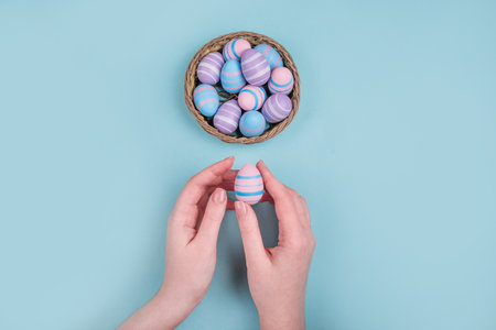 Woman hands holding dyed on blue, pink and violet Easter eggs in the straw basket isolated on blue...