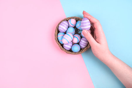 Woman hand holding dyed on blue, pink and violet Easter eggs in the straw basket isolated on blue background. Easter theme flatlay.の写真素材