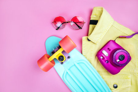 Summer fashion flatlay with bright yellow denim jacket, turquoise skateboard, round gradient sunglasses and instant camera isolated on pink background.の写真素材