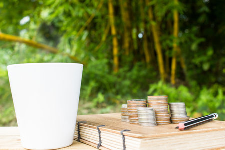 Coins, notebook and white cup on wooden table on green nature background. Conceptual business and relaxing in nature.の写真素材