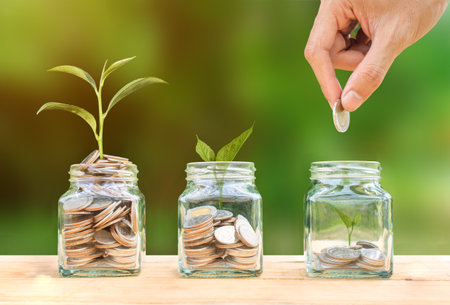 Conceptual hand putting coins into glass bottle and growing tree on nature background,Business investment growth conceptの写真素材