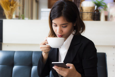 A beautiful asian business woman sitting on sofa drinking cup of hot coffee and using smart phone to check meeting, email or message today.  Business woman relaxing in coffee shop. Copy space.の写真素材