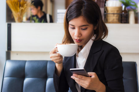 Asian business woman relaxing in coffee shop with hot coffee. Copy space. Coffee is a relaxing place for everyone today. Hand holding white cup of coffee and checking e-mail or message today.の写真素材