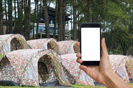 Hand holding black smartphone with white screen for mock up on camping tent as background.