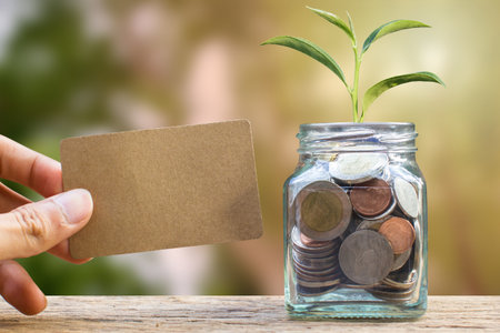 Saving and invest money concept. Conceptual hand holding brown card near the glass jar on nature...