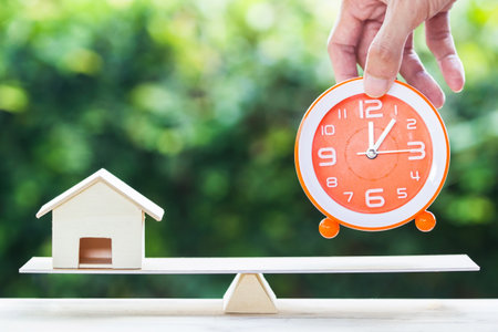 Home bargain, real estate, loan, house lender reverse mortgage concept. Balance orange clock and hand holding small residence on wooden table and blurred green nature as background. Time and home.の写真素材