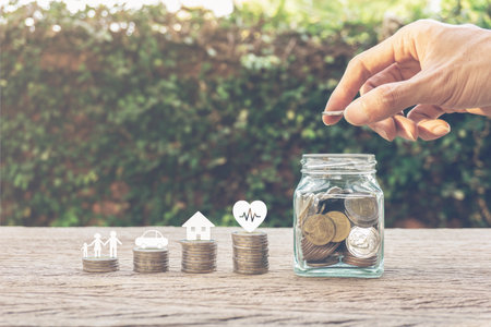 Savings money for family life concepts. Hand holding coin on a full money in glass jar and family member, car, house, healthy on coins. Depicts saving for wealth and life. fundraising concept.の写真素材