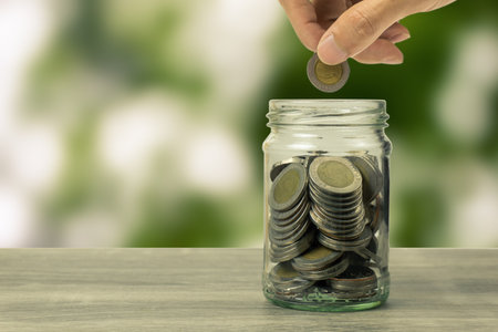 Savings and investment concept. Hand putting coin in glass jar on table on green nature background....