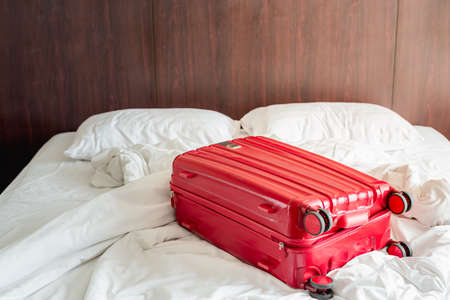 Messy and unmade white traveller's hotel bed at hotel room with red luggage.の写真素材