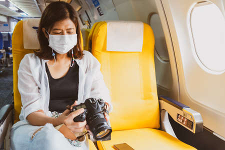 Asian woman traveler wearing face mask looking photography from her trip in dslr camera on board in airplane.の写真素材