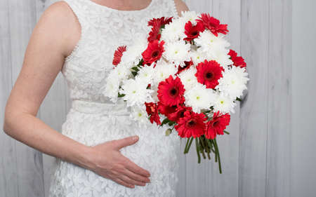 A pregnant woman over 40 in a white dress holds her belly with her hand, a bouquet of white and red flowers in the other hand, copy spaceの写真素材