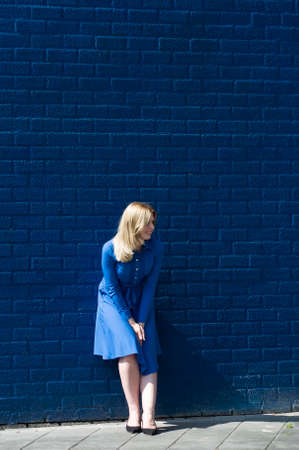 blonde with flying hair in a blue dress stands by a blue brick wall on a sunny day and laughsの写真素材