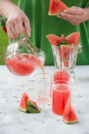 young woman in a green dress pours a red refreshing drink from watermelon pulp into a glass glass, bright natural backgroundの写真素材