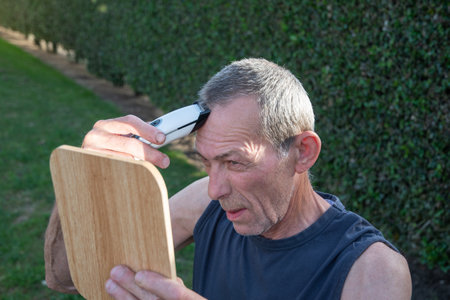 a gray-haired middle-aged man shaves his hair with a clipper in a garden