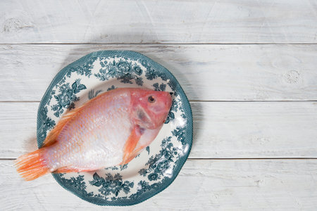 raw pink tilapia fish lies on a plate with blue ornaments on white wooden boardsの写真素材