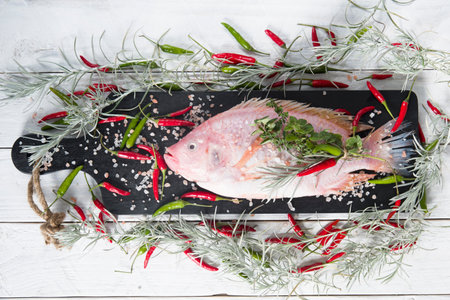 raw fresh pink tilapia fish lies on a board sprinkled with small red hot peppers