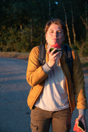 Young Caucasian blonde millennial woman looking like a guy eating watermelon in the sunny golden hour at sunsetの写真素材