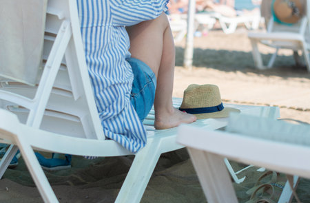 Young woman sitting on a sun lounger on the beach next to a straw hat, woman looking pensive,...
