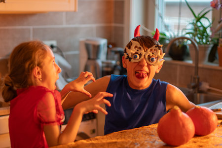 brother and sister showing scary faces while decorating house with Halloween decor, girl in devil costume, teenage boy in ghost glasses playing in darkened room, traditional autumn holidayの写真素材