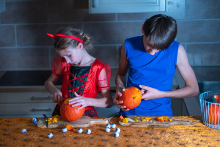 Little girl in a Halloween costume and her brother cut out eyes on a pumpkin with a sharp knife in a darkened room, a traditional autumn holiday.の写真素材