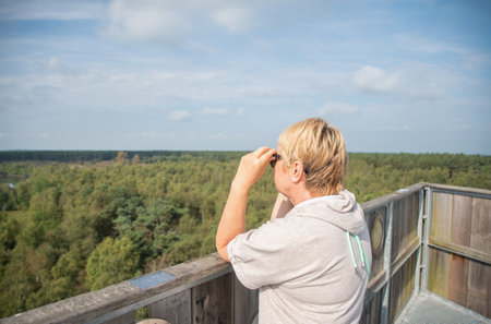 middle aged blonde woman with short haircut gazes into the distance at the landscape from an observation tower on beautiful lake in Belgium, sunny autumn dayの写真素材