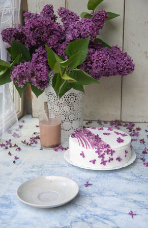 delicate still life with a cut cake and a glass of coffee with milk, decorated with lilac flowers, near a vase with lilac branches on a wooden table, emphasizing the floral and rustic lifestyle,の写真素材