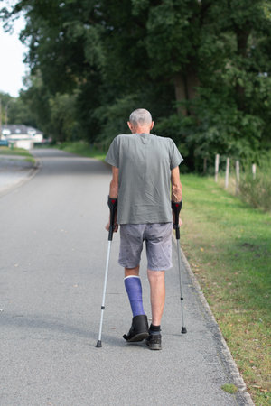 A man with a broken leg is walking down the street, on his left leg he has a special boot for walking, if the leg is in a cast, the man moves with the help of crutches,high quality photoの写真素材