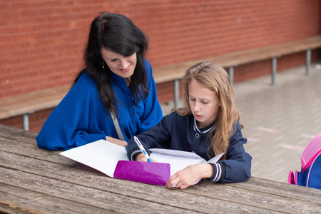 loving mother watching her daughter doing homework on her own at wooden table in childrens playground, education concept, back to school, girl in beautiful blue uniform,の写真素材