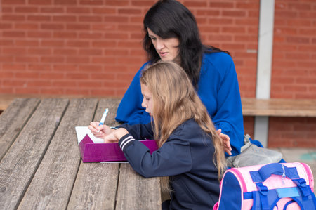 young mother lovingly watches her daughter doing homework at a wooden table during a walk in the park outdoors, they smile happily together, back to school,の写真素材