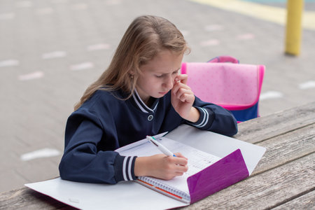 Teenage girl focused on solving complex math equation at wooden table in park outdoors, education concept, back to school, girl in beautiful blue uniform and with purple notebookの写真素材